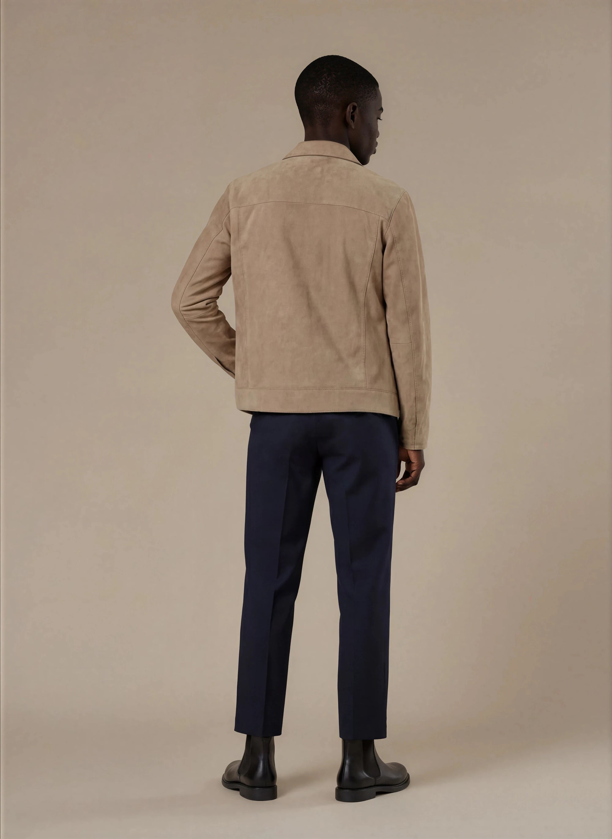 Back view of man wearing a beige suede jacket and dark formal pants with black chelsea boots